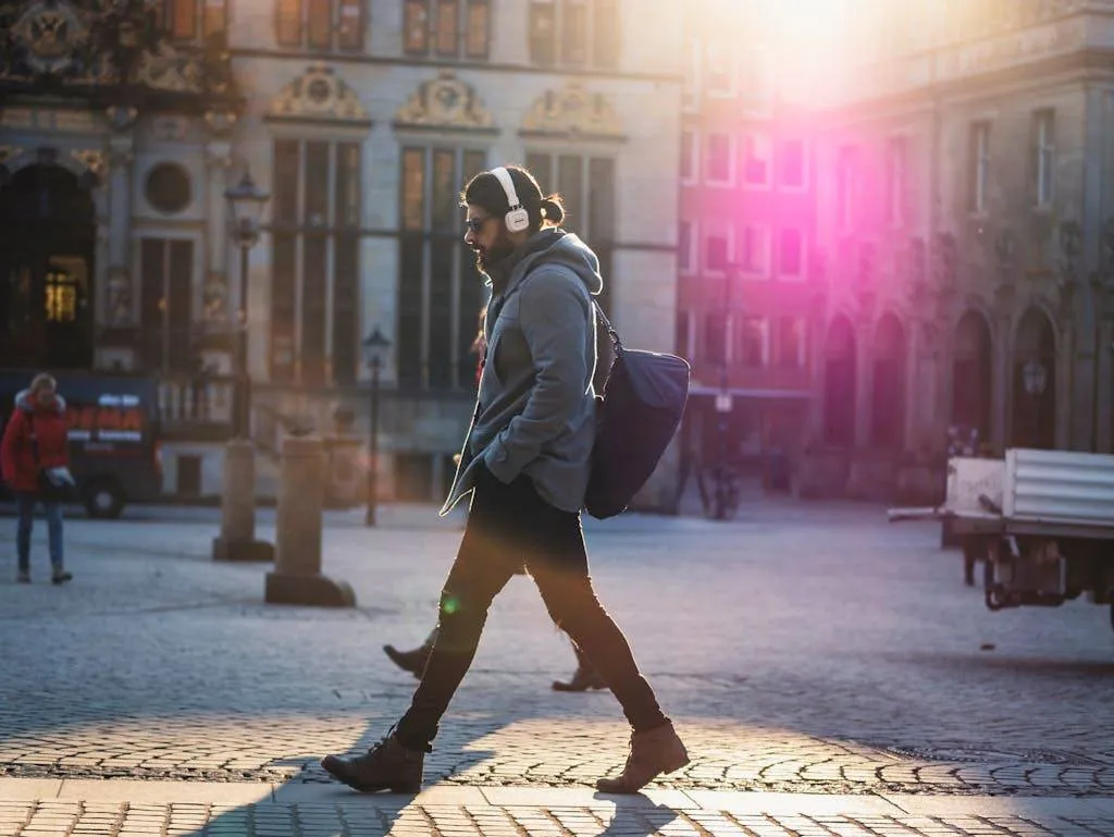 Man with headphones walks down a sunlit urban street, casting a shadow on the cobblestones.