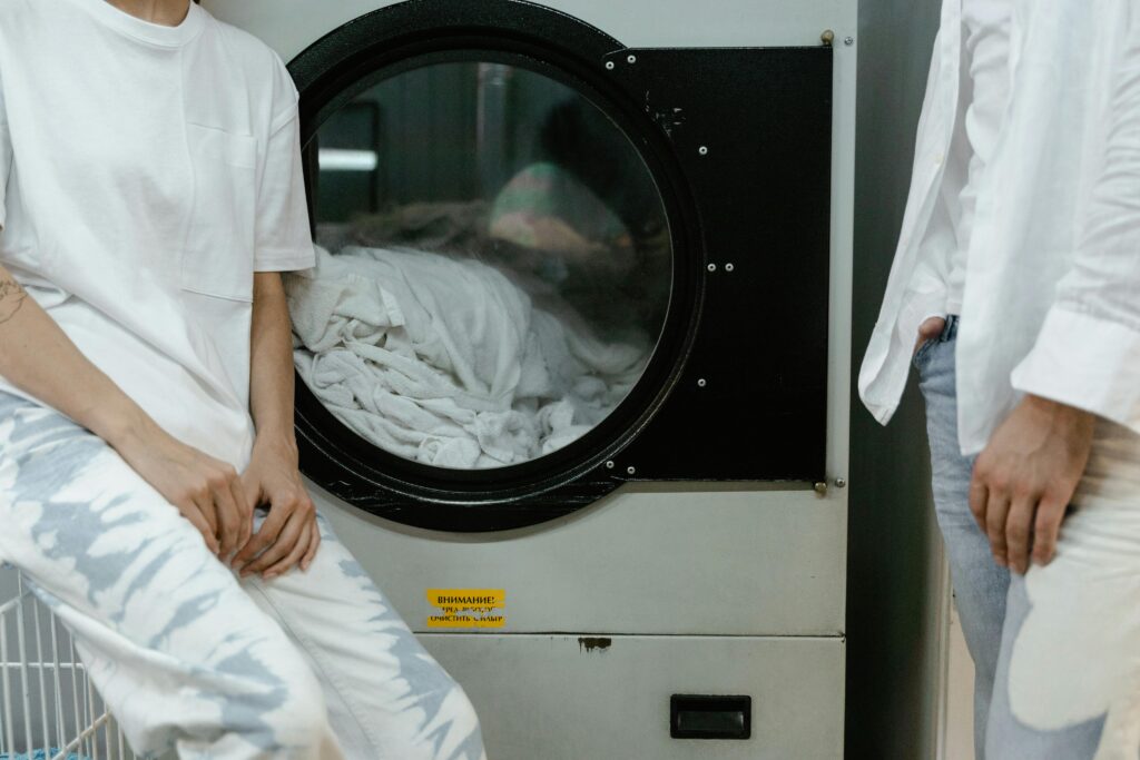 Adults in white clothes using a washing machine in a laundry facility.